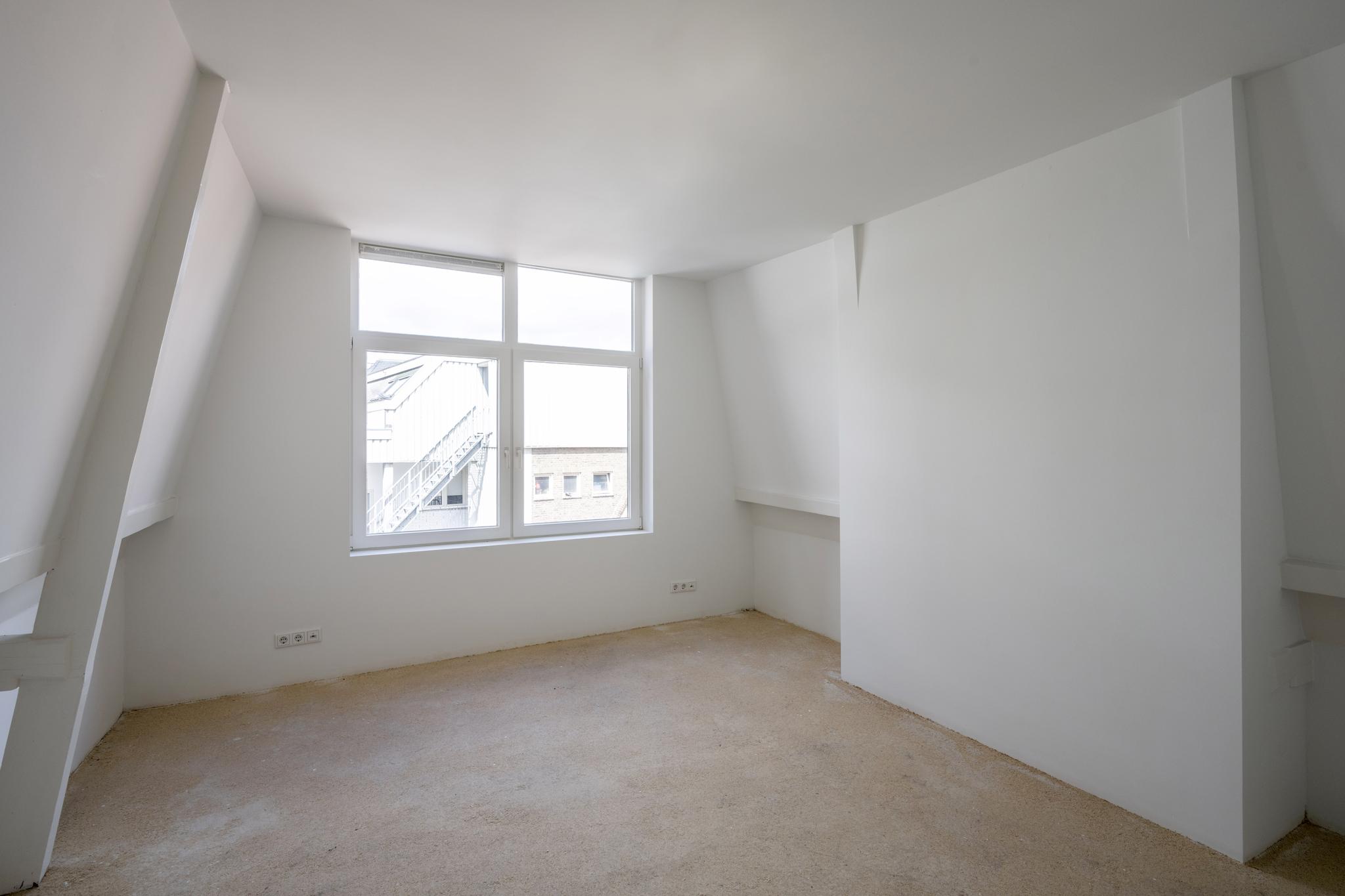 Lichte, lege zolderkamer met witgeschilderde schuine muren en een groot raam met uitzicht op gebouwen aan het Damrak.
