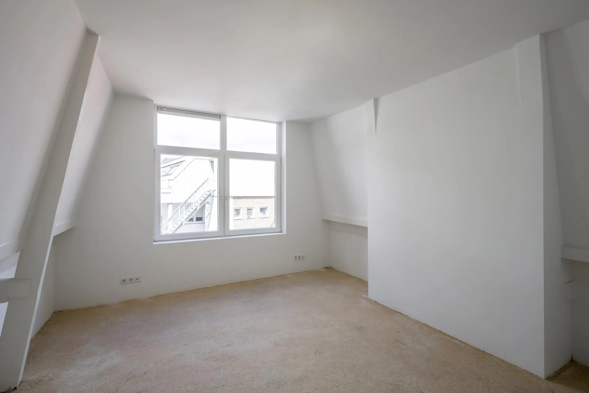 Lichte, lege zolderkamer met witgeschilderde schuine muren en een groot raam met uitzicht op gebouwen aan het Damrak.