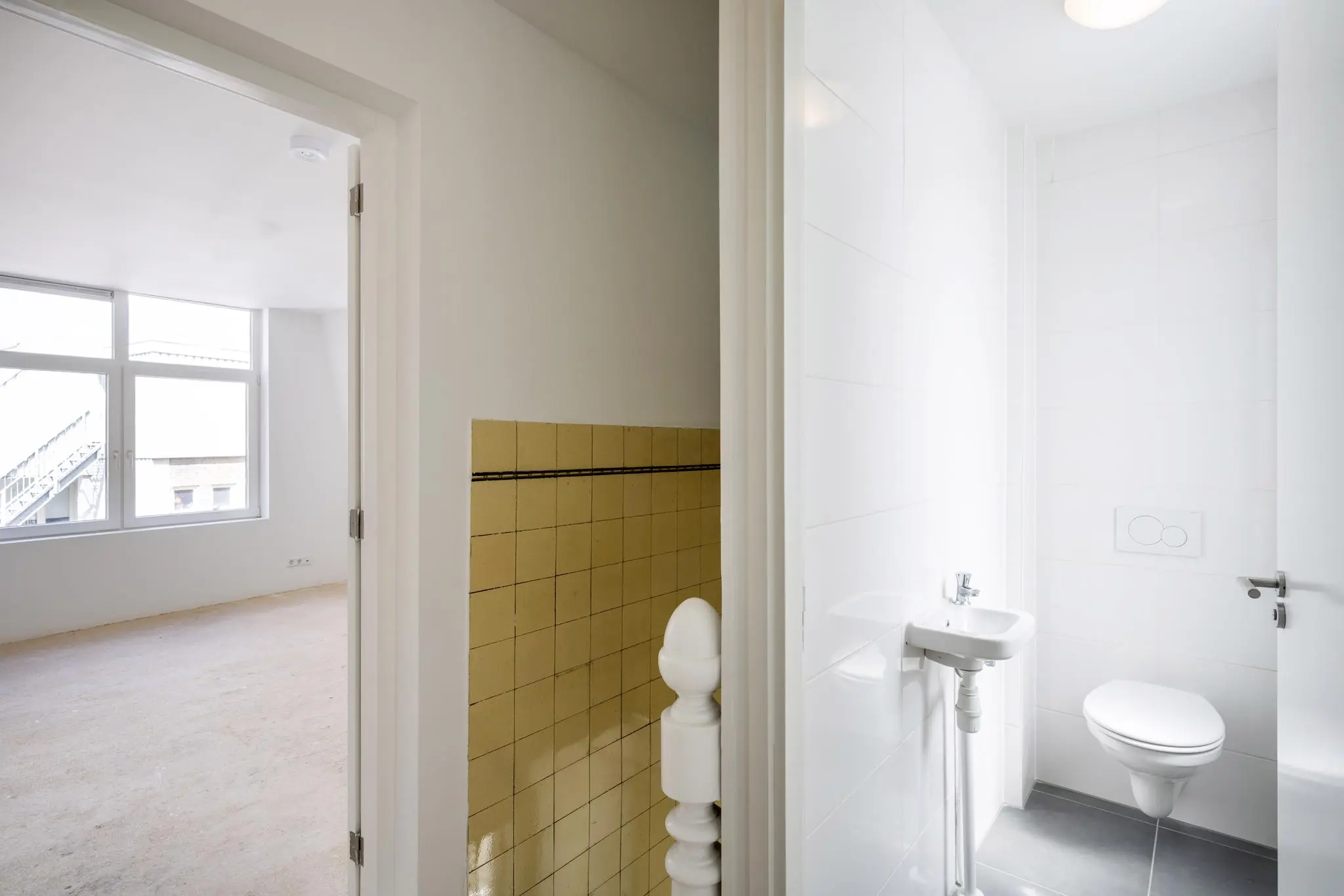 Wit betegeld toilet met wandcloset en fonteintje naast een lichte kamer met groot raam aan het Damrak.