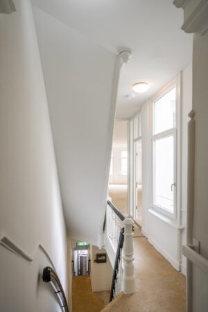 Trappenhuis met witgeschilderde muren, houten leuning en grote ramen in een pand aan het Damrak.