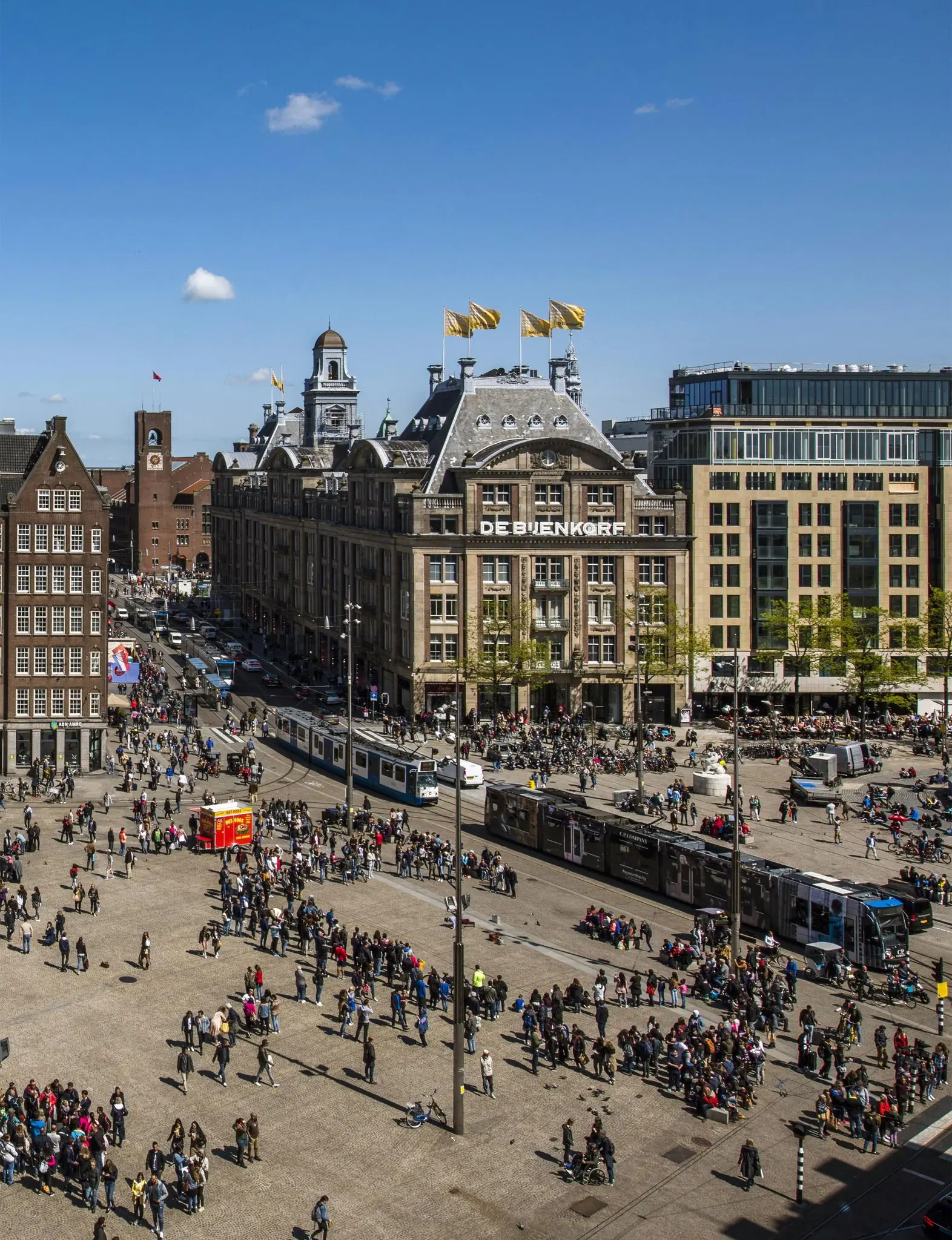 Drukke Damrak in Amsterdam met winkelgebouw De Bijenkorf, trams en veel voetgangers op het plein.