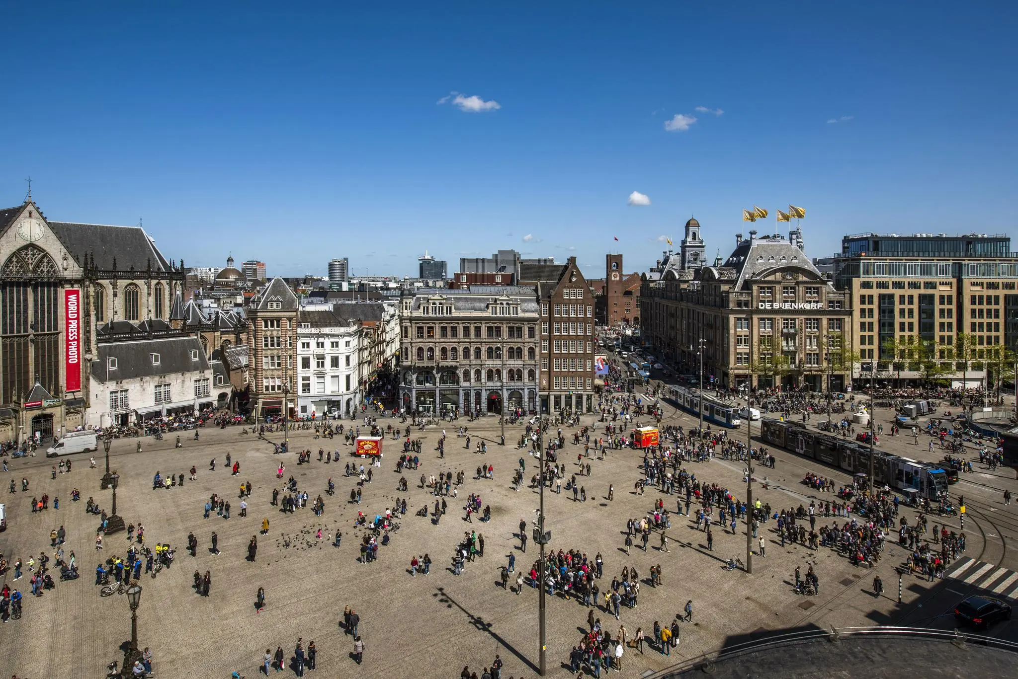 Druk Damrak met uitzicht op de Dam in Amsterdam, omringd door historische gebouwen en bezoekers onder een blauwe lucht.