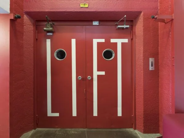 Rode liftdeuren met het woord "LIFT" in witte letters in een rood trappenhuis aan de Generaal Vetterstraat.
