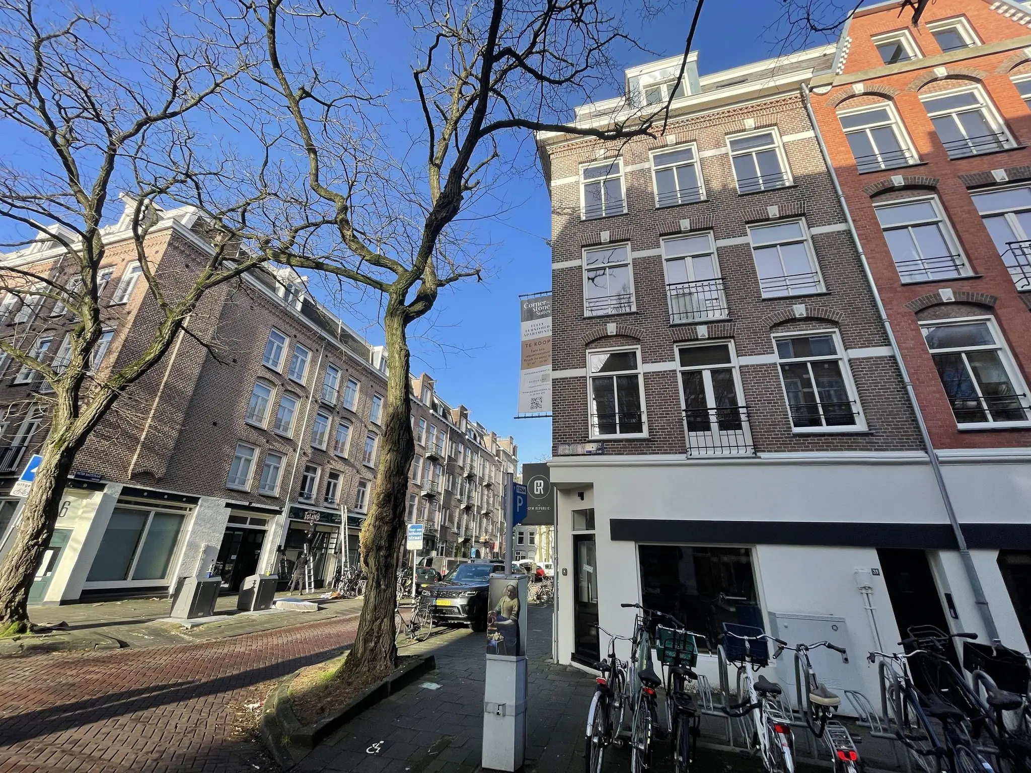 Straatbeeld van de Pieter Baststraat in Amsterdam met bakstenen gebouwen, geparkeerde fietsen en kale bomen onder een blauwe lucht.