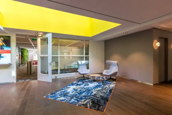 Moderne wachtruimte in kantoorpand aan de Newtonlaan met twee designstoelen, kleurrijk vloerkleed en groot geel plafondraam.