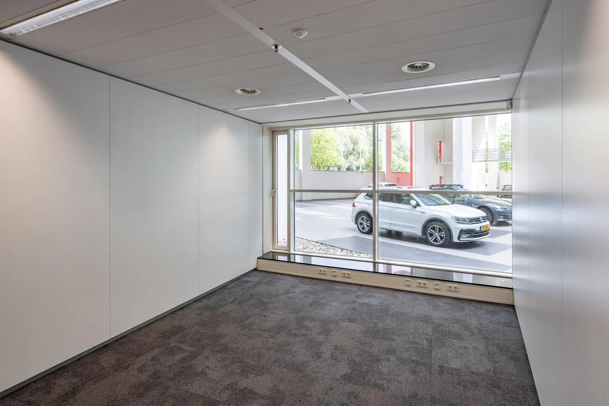 Leeg kantoor met tapijtvloer en groot raam met uitzicht op geparkeerde auto's aan de Newtonlaan.