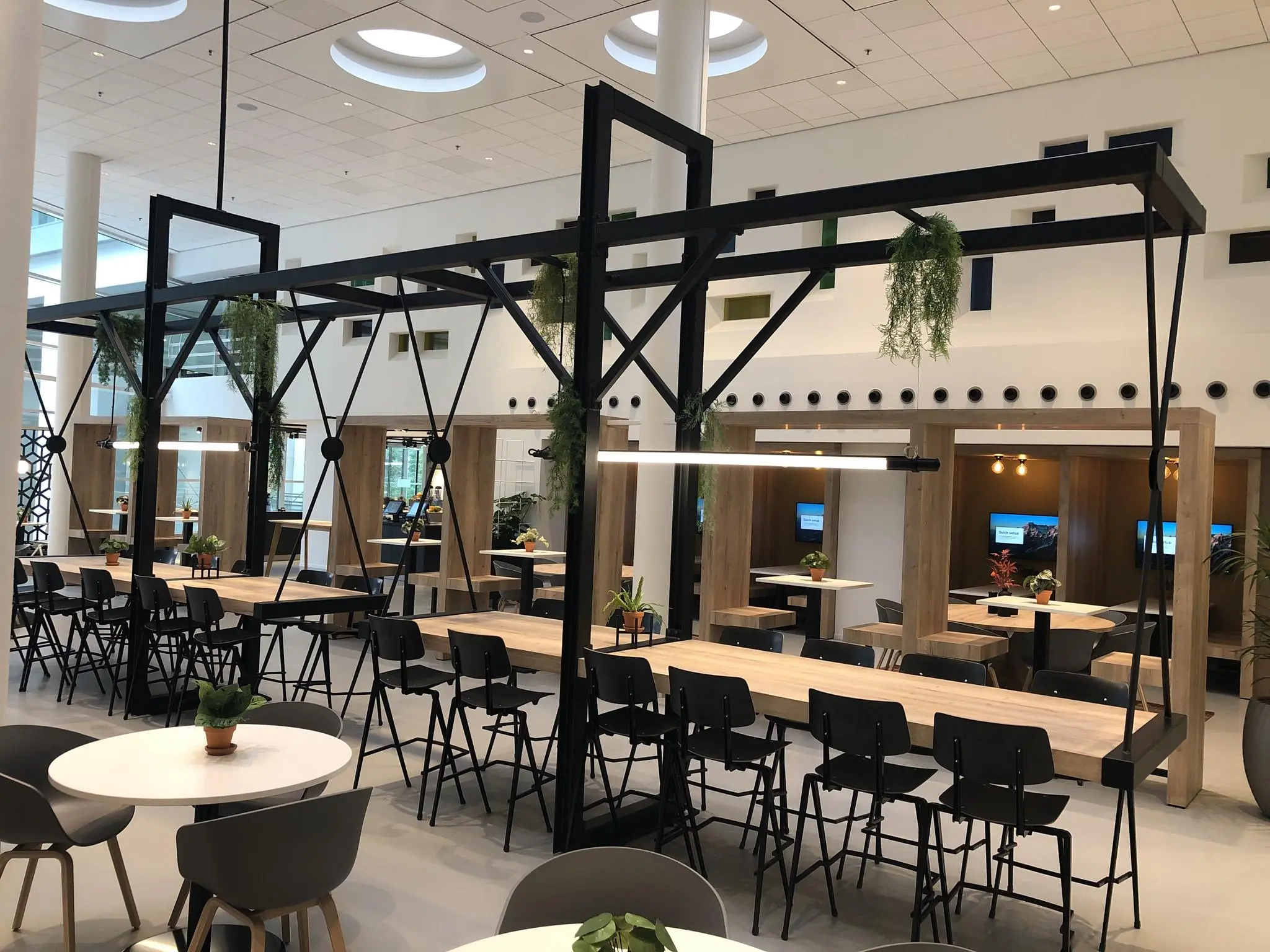 Moderne en open kantoorkantine aan de Newtonlaan met lange houten tafels, hangplanten en werkplekken met schermen.