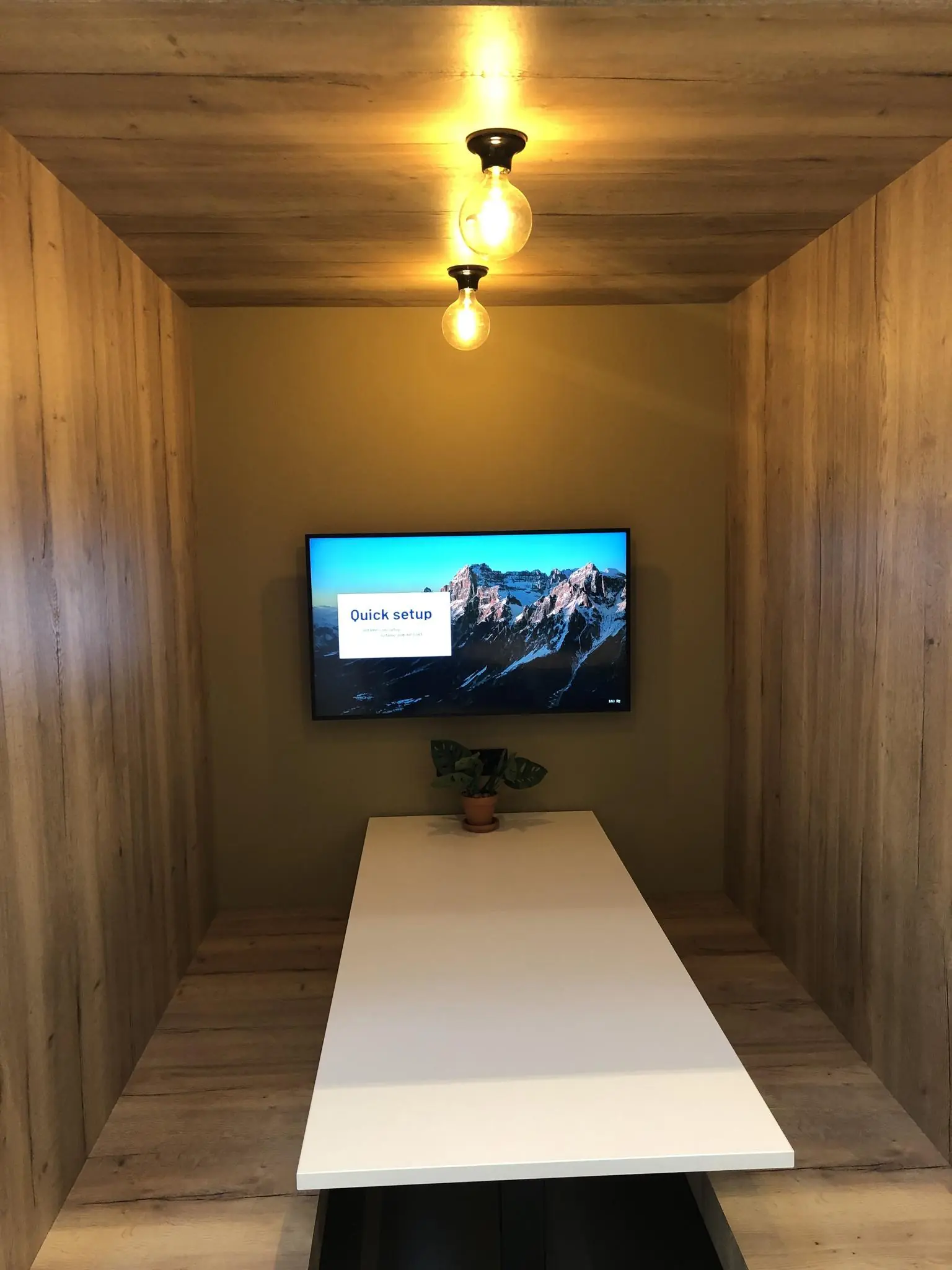 Vergaderruimte met houten wanden, witte tafel, een beeldscherm met opschrift "Quick setup" en een plantje als decoratie.