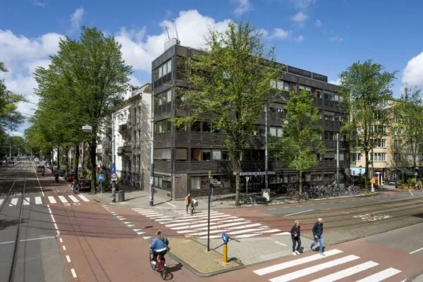 Straatbeeld van de Sarphatistraat in Amsterdam met een modern hoekgebouw, fietsers, voetgangers en trambaan.