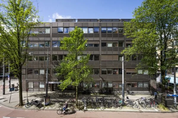 Vooraanzicht van een modern kantoorgebouw aan de Sarphatistraat in Amsterdam met fietsen geparkeerd op de stoep en bomen langs de straat.