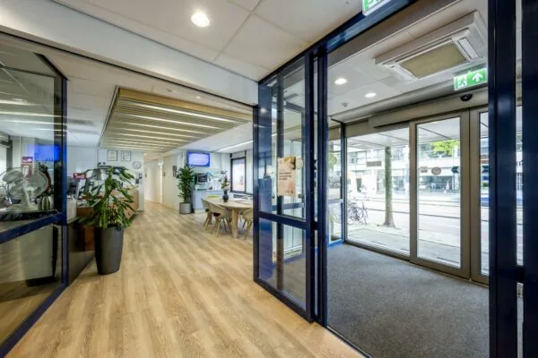Ingang en ontvangstruimte van een modern kantoorpand aan de Sarphatistraat met houten vloer, glazen deuren en wachtruimte.