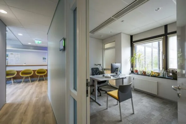 Moderne spreekkamer aan de Sarphatistraat met bureau, computer en uitzicht op wachtruimte met gele stoelen.