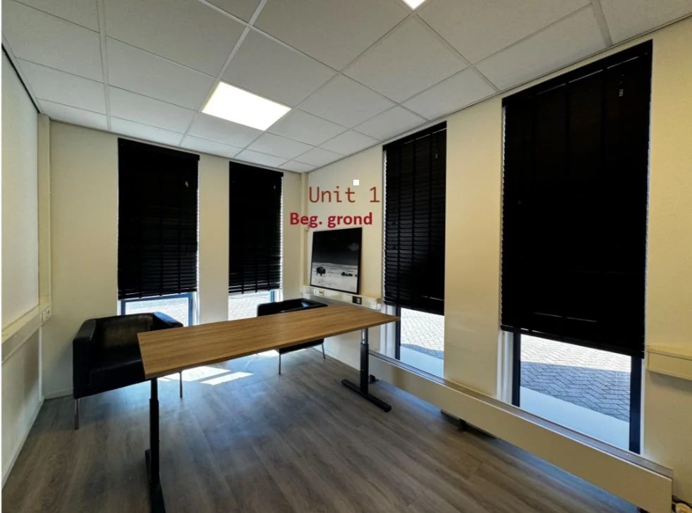 Kantoorruimte Unit 1 op de begane grond met houten bureau, zwarte stoelen en ramen met zwarte jaloezieën.