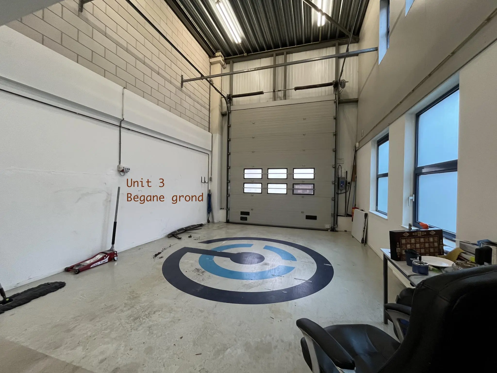 Interieur van een bedrijfsruimte met grote garagedeur, betonnen vloer en de tekst "Unit 3 Begane grond" op de muur.