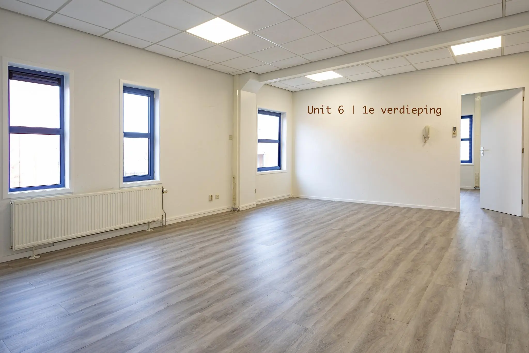 Lichte, lege kantoorruimte met drie ramen, grijze vloer en de tekst "Unit 6 | 1e verdieping" op de muur.