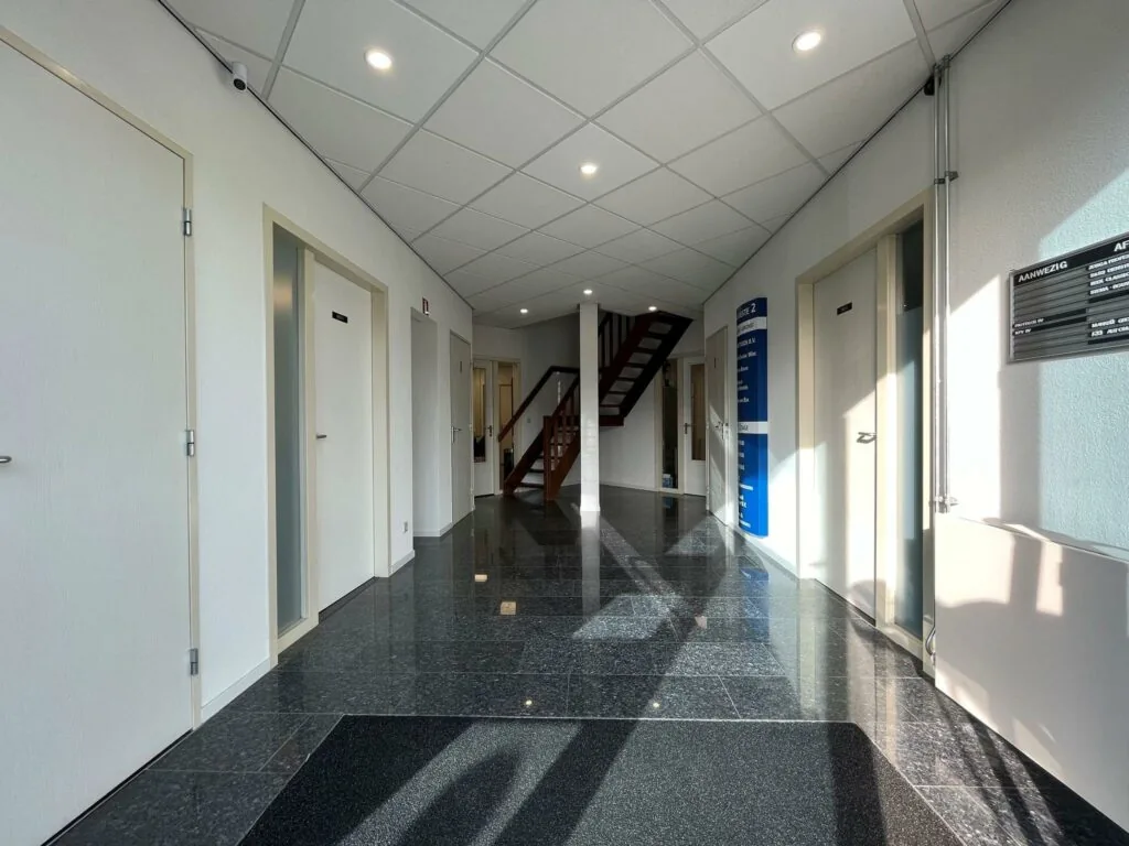 Ingangshal van kantoorpand Bergveste met witte deuren, glanzende zwarte vloer en een houten trap in het midden.