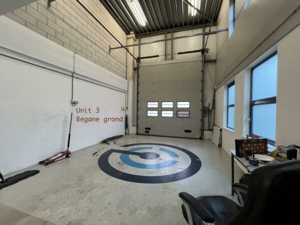 Interieur van werkruimte Unit 3 op de begane grond met industriële garagedeur en bedrijfslogo op de vloer.