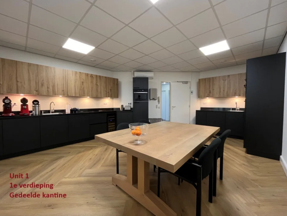 Moderne gedeelde kantine op de eerste verdieping van Unit 1 met een grote houten tafel, zwarte keukenkasten en houtkleurige bovenkasten.