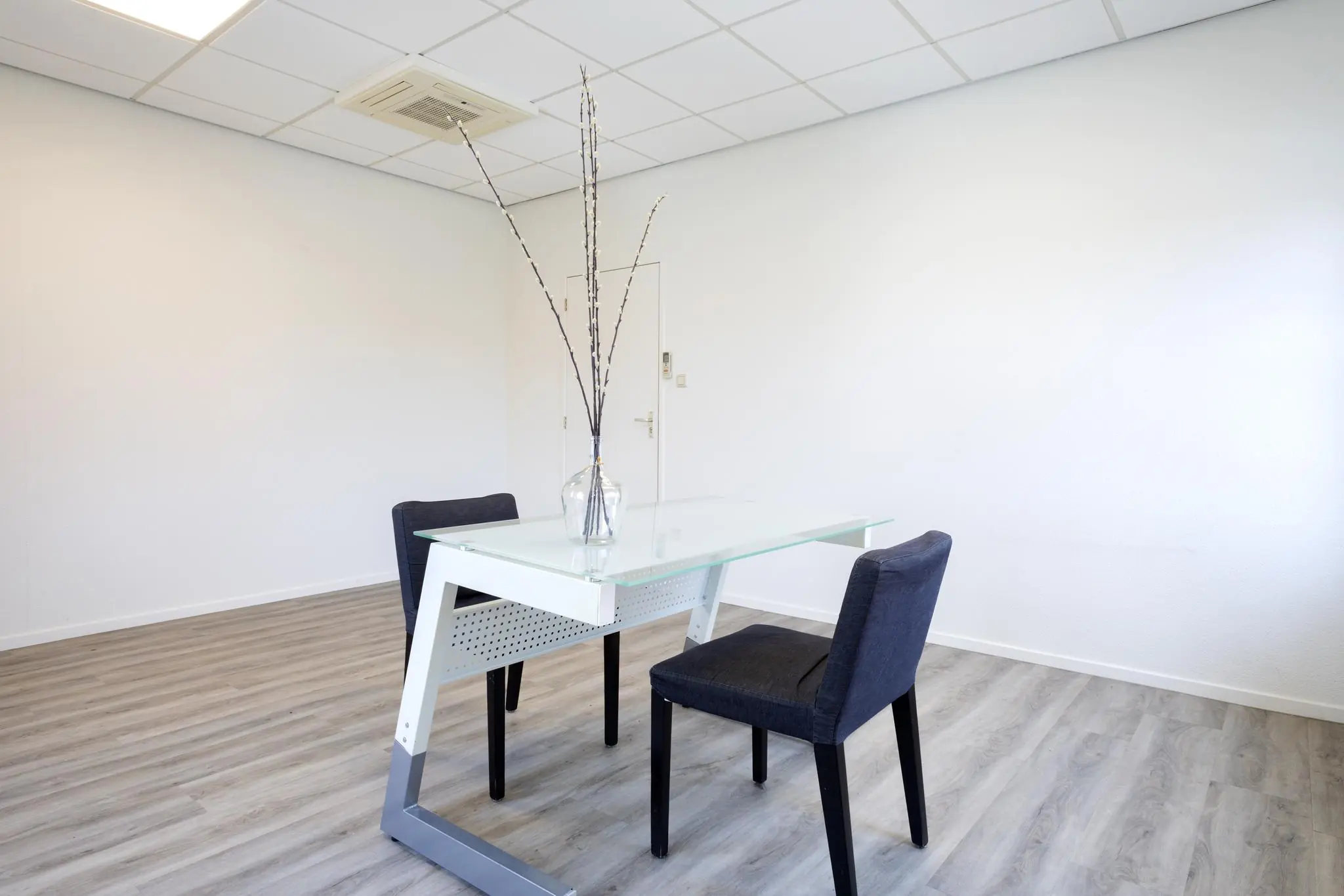Minimalistisch ingericht kantoor met twee stoelen, een glazen bureau en een vaas met takken.