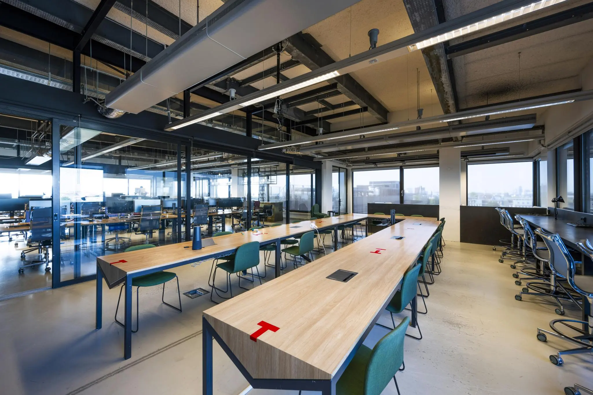 Moderne, open kantoorruimte aan de Vijzelstraat met lange houten bureaus, groene stoelen en glazen scheidingswanden.