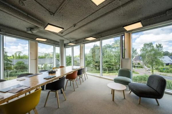 Modern vergaderkamerinterieur met lange houten tafel, kleurrijke stoelen en grote ramen met uitzicht op het John M. Keynesplein.