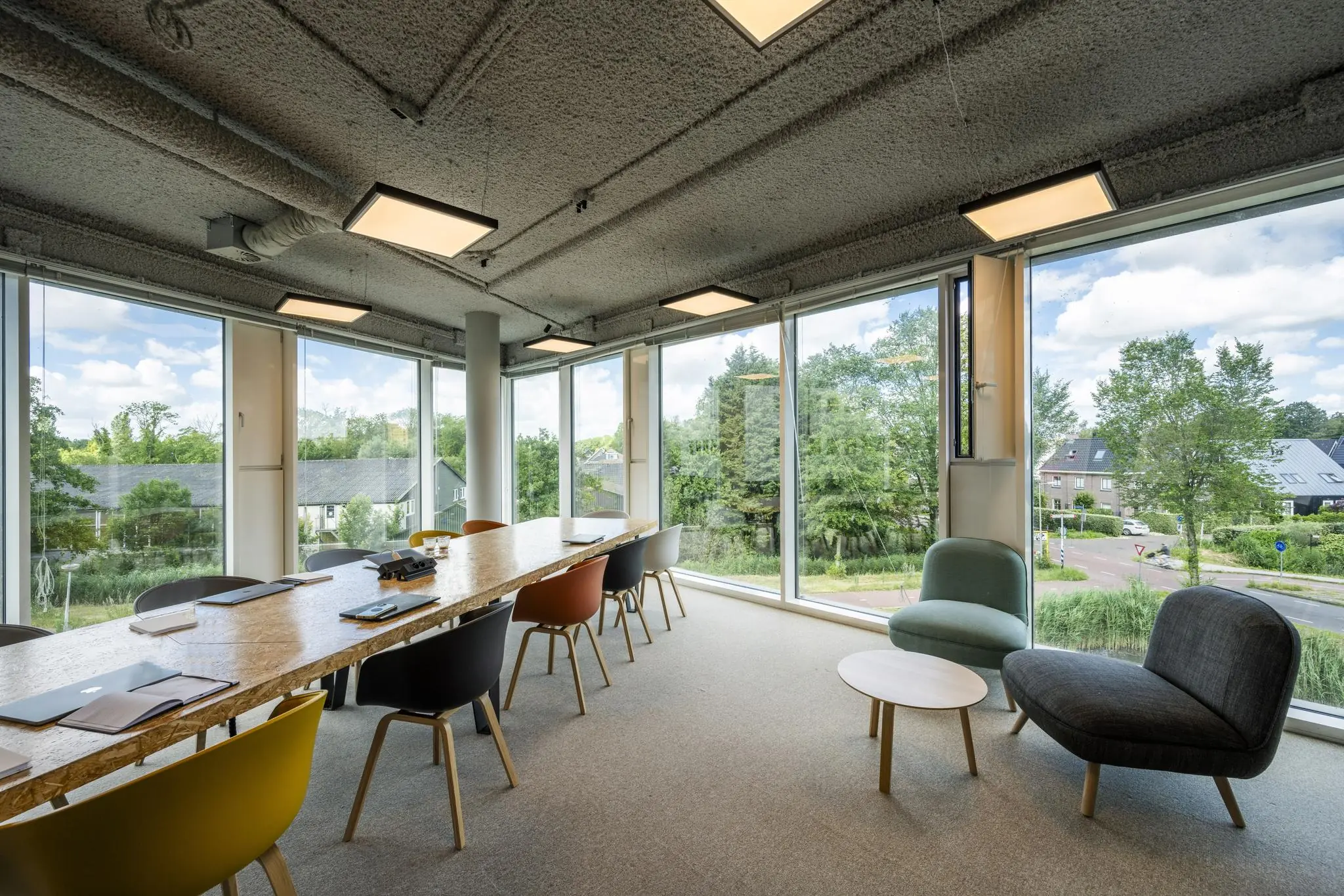 Modern vergaderkamerinterieur met lange houten tafel, kleurrijke stoelen en grote ramen met uitzicht op het John M. Keynesplein.