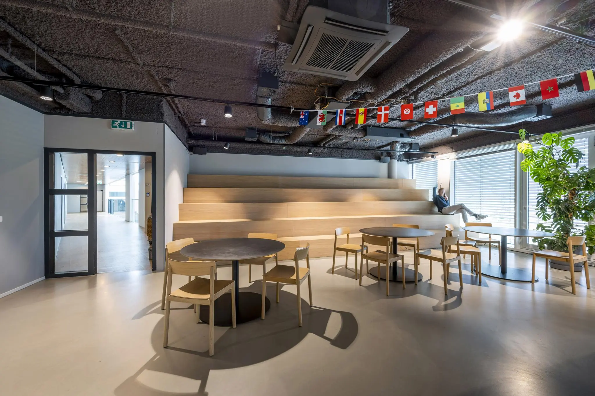 Moderne kantineruimte aan het John M. Keynesplein met ronde tafels, houten tribunezitplaatsen en internationale vlaggen aan het plafond.