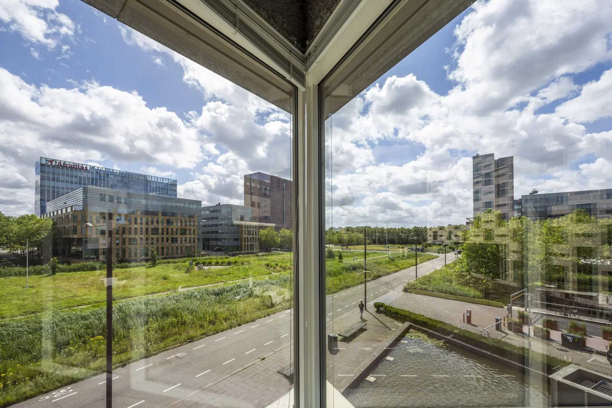Uitzicht vanuit een raam op moderne kantoorgebouwen aan het John M. Keynesplein in Amsterdam met een groen landschap en heldere lucht.