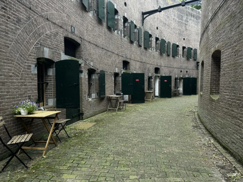 Binnenplaats van Fort bij Vechten aan de Lekdijk, met groene deuren, bakstenen muren en houten tafeltjes met bloemen.