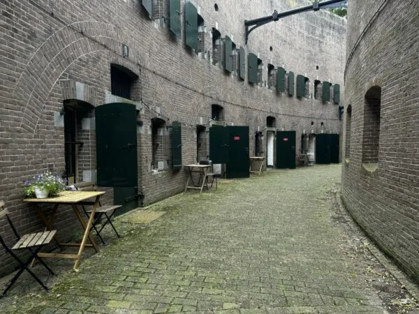 Binnenplaats van Fort bij Vechten aan de Lekdijk, met groene deuren, bakstenen muren en houten tafeltjes met bloemen.