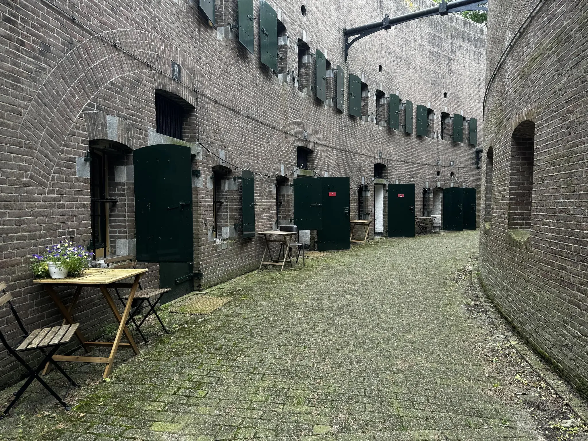 Binnenplaats van Fort bij Vechten aan de Lekdijk, met groene deuren, bakstenen muren en houten tafeltjes met bloemen.