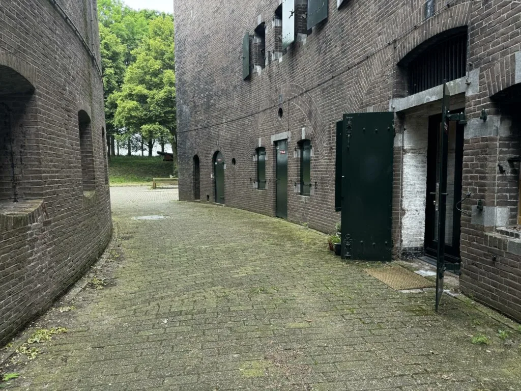 Binnenplaats tussen twee bakstenen gebouwen van Fort bij Vreeswijk aan de Lekdijk, met openstaande groene deuren en zicht op bomen op de achtergrond.