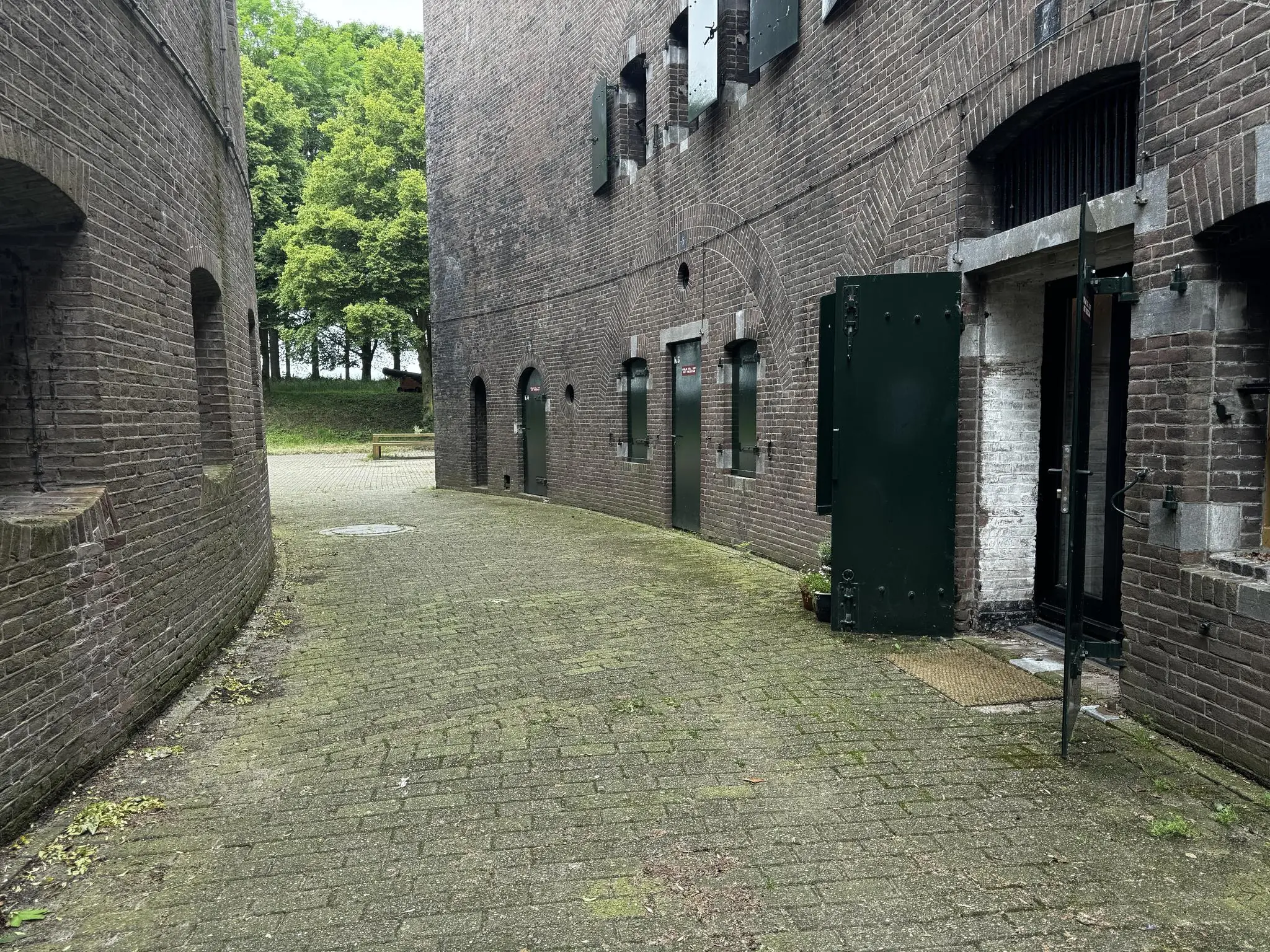 Binnenplaats tussen twee bakstenen gebouwen van Fort bij Vreeswijk aan de Lekdijk, met openstaande groene deuren en zicht op bomen op de achtergrond.