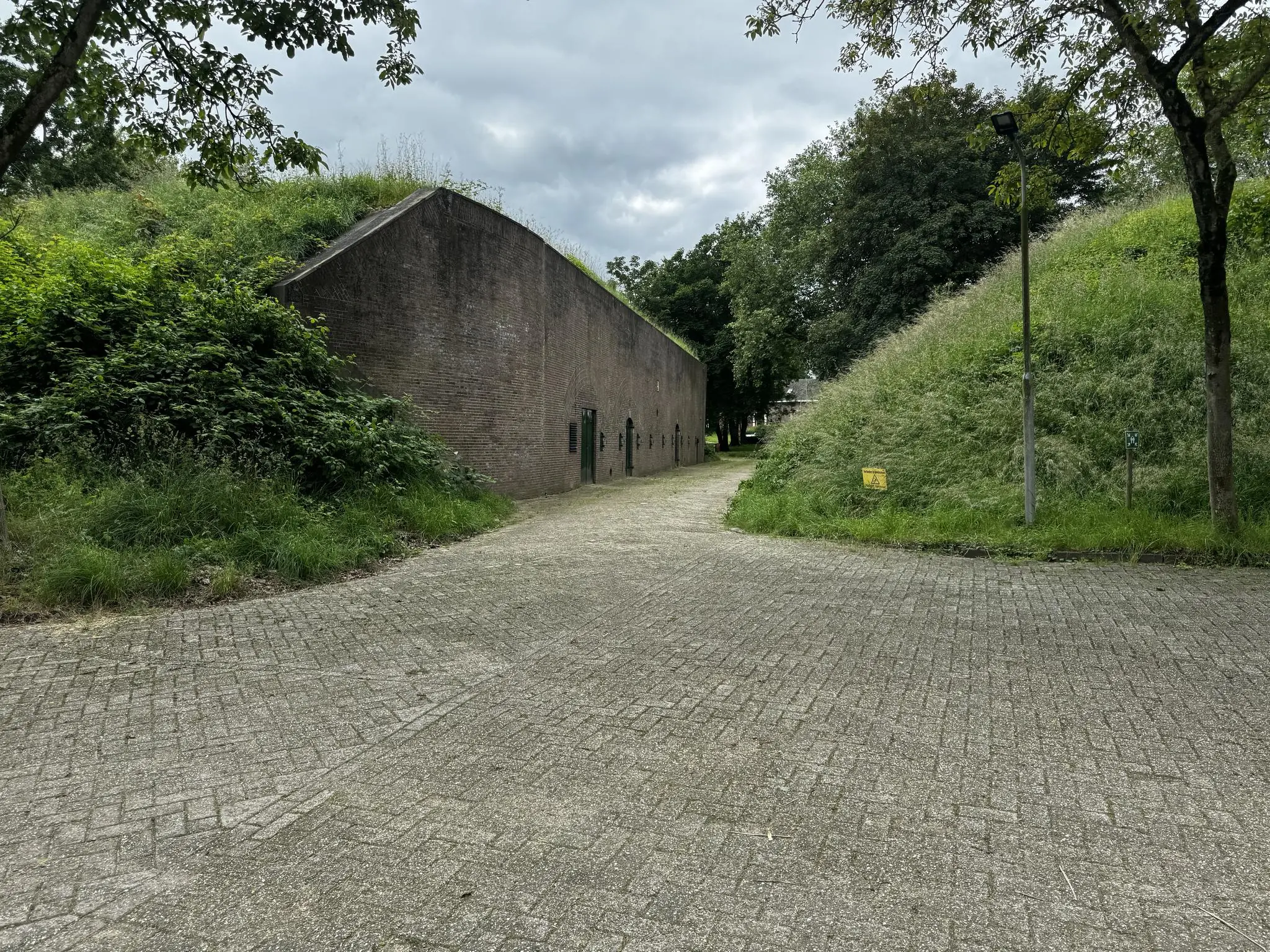 Een met gras begroeid verdedigingswerk aan de Lekdijk met een smal pad ertussen en omringd door bomen.