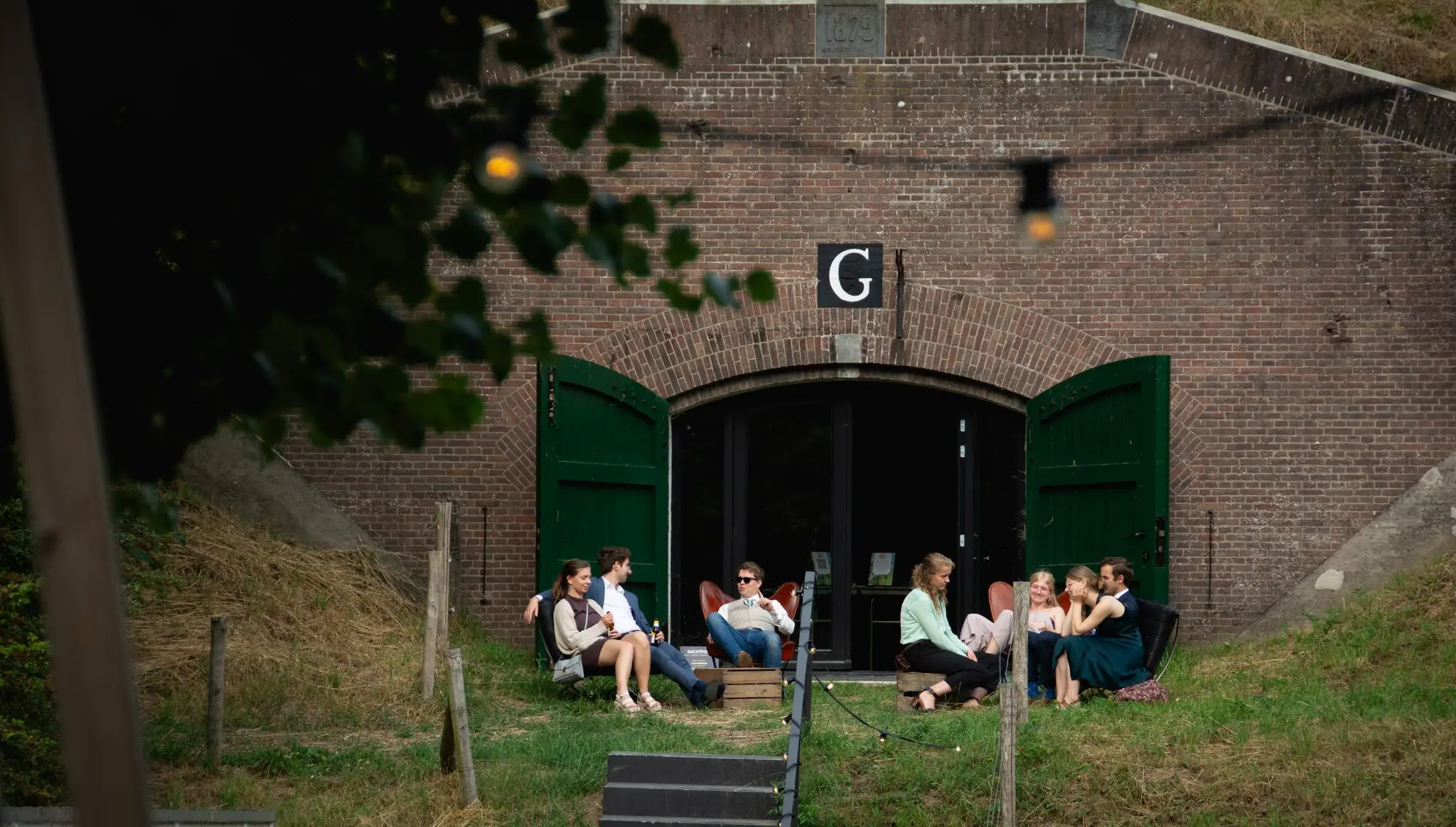 Groep mensen zit ontspannen op stoelen voor een openstaande deur van een vestinggebouw aan de Lekdijk.