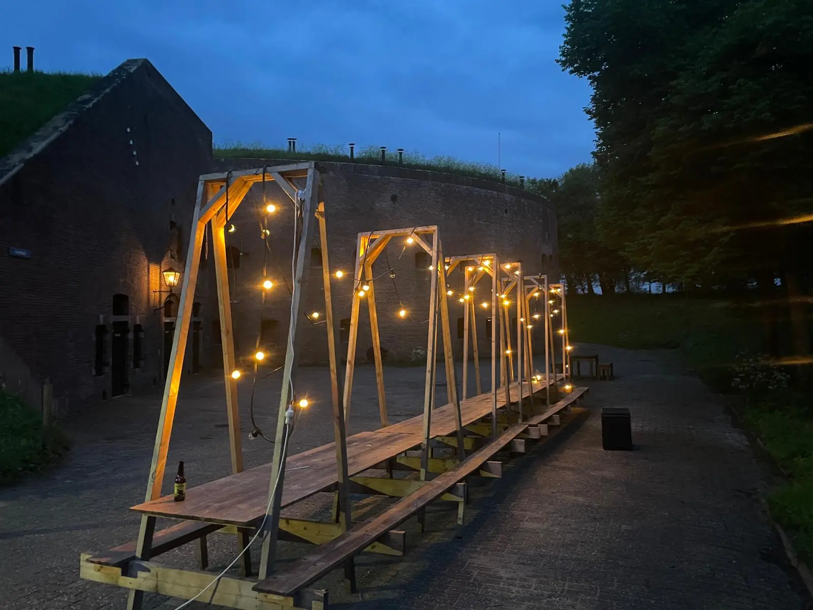 Lange houten tafel met lichtslingers bij schemering voor een historisch fort aan de Lekdijk.