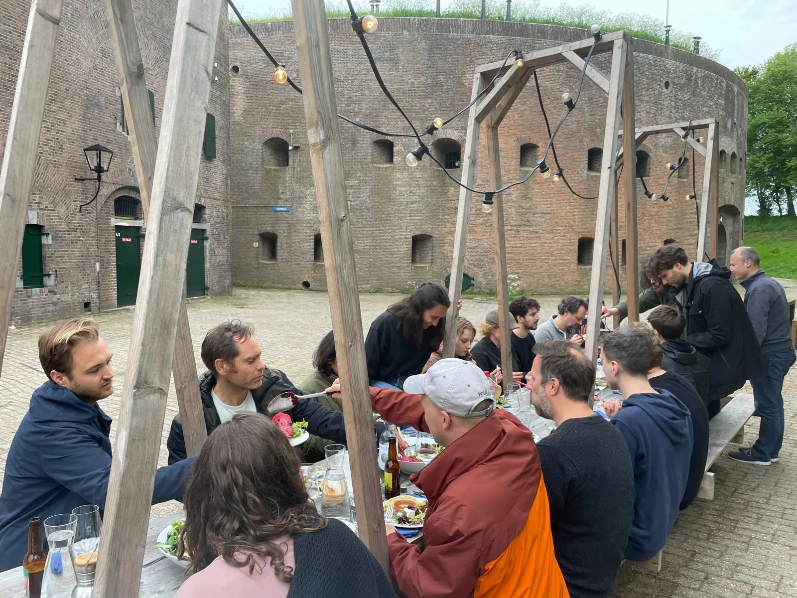 Een groep mensen eet samen aan een lange picknicktafel in de buitenlucht bij Fort bij Vechten aan de Lekdijk.