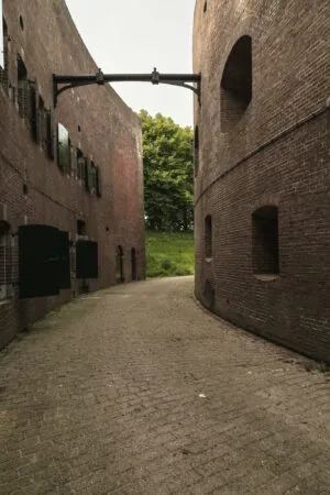 Smalle geklinkerde doorgang tussen twee bakstenen vestinggebouwen bij Lekdijk, met openstaande luiken en uitzicht op groene bomen.