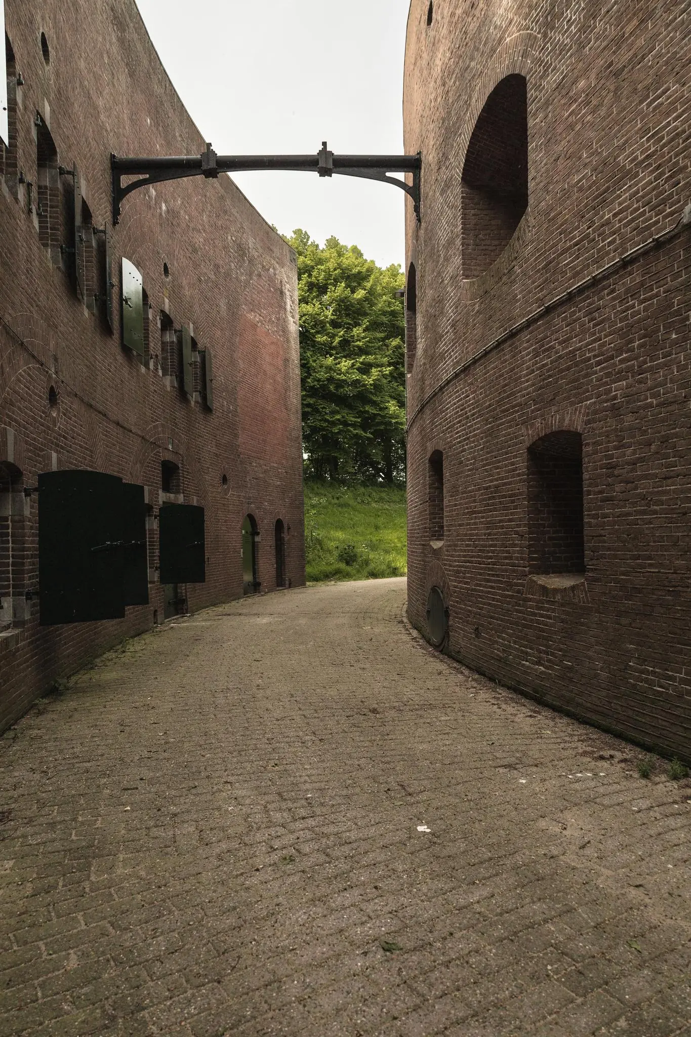 Smalle geklinkerde doorgang tussen twee bakstenen vestinggebouwen bij Lekdijk, met openstaande luiken en uitzicht op groene bomen.