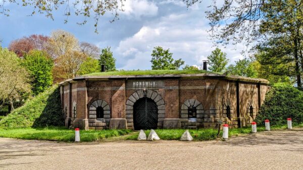 Rond fortgebouw van baksteen met grasdak, genaamd Fort Honswijk, omgeven door bomen en een verharde weg.