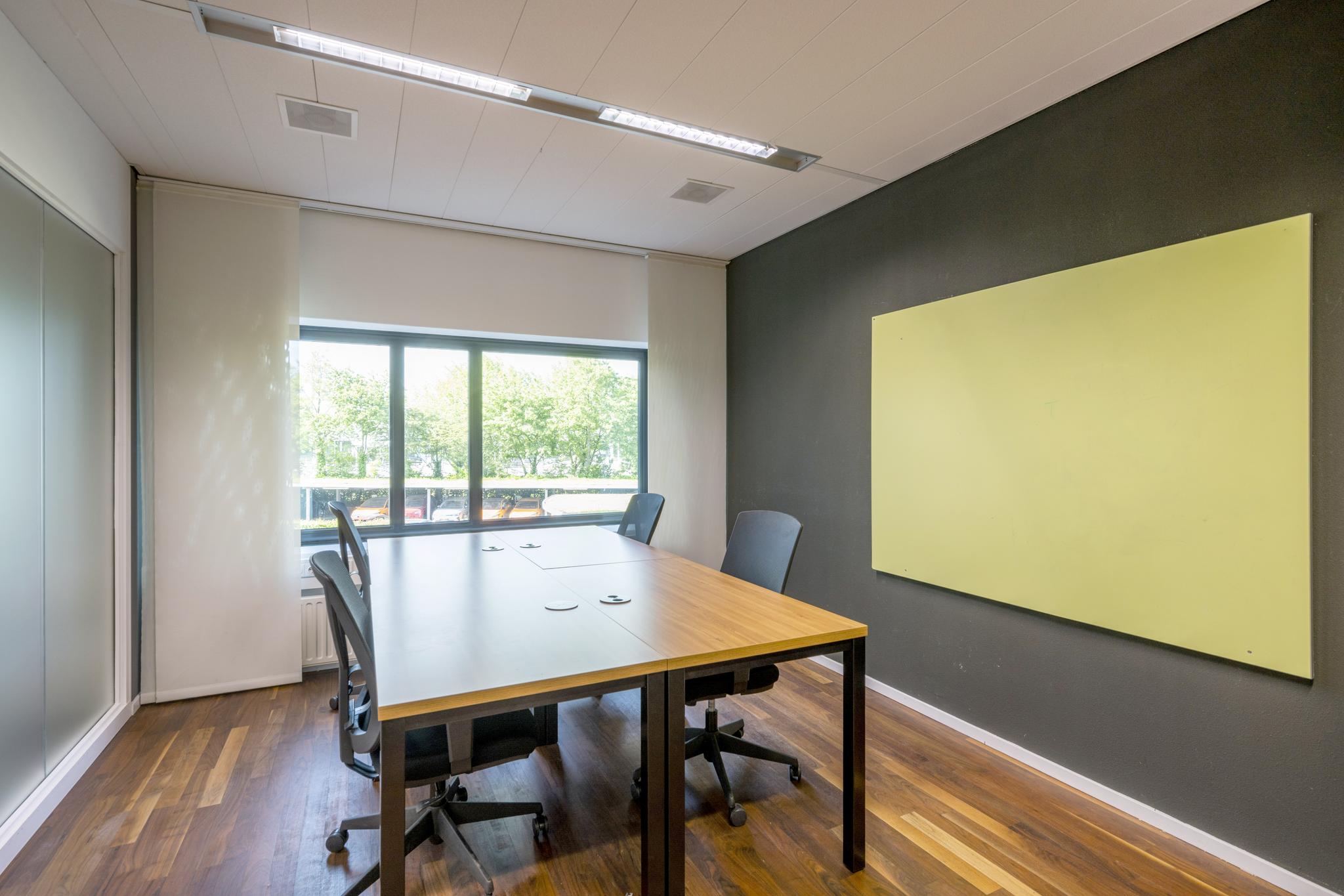 Vergaderruimte met houten tafel, vier bureaustoelen, groot raam en een lichtgele wandplaat aan een donkere muur.