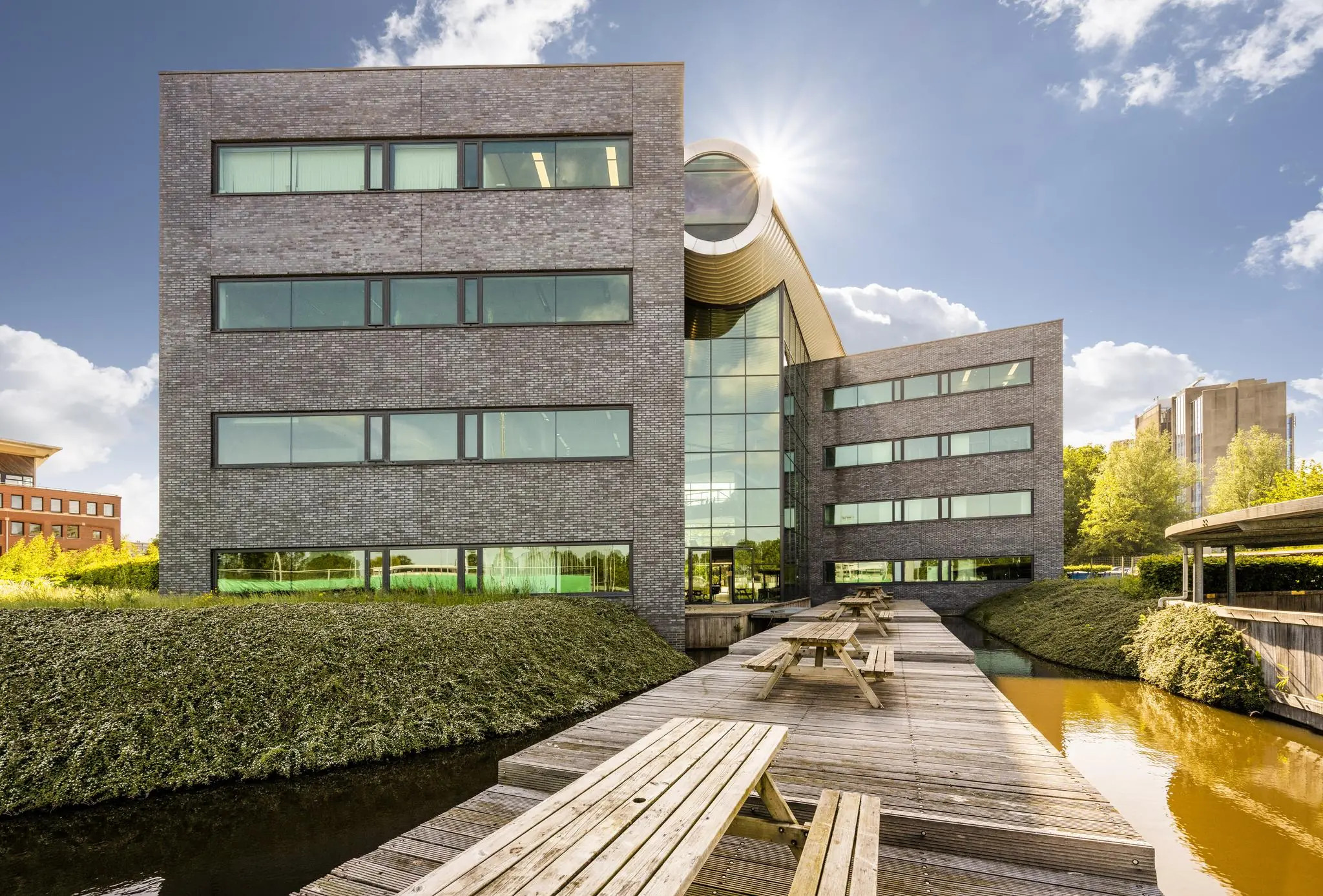 Modern kantoorgebouw aan de Prof. W.H. Keesomlaan met houten picknicktafels op een vlonder boven het water.