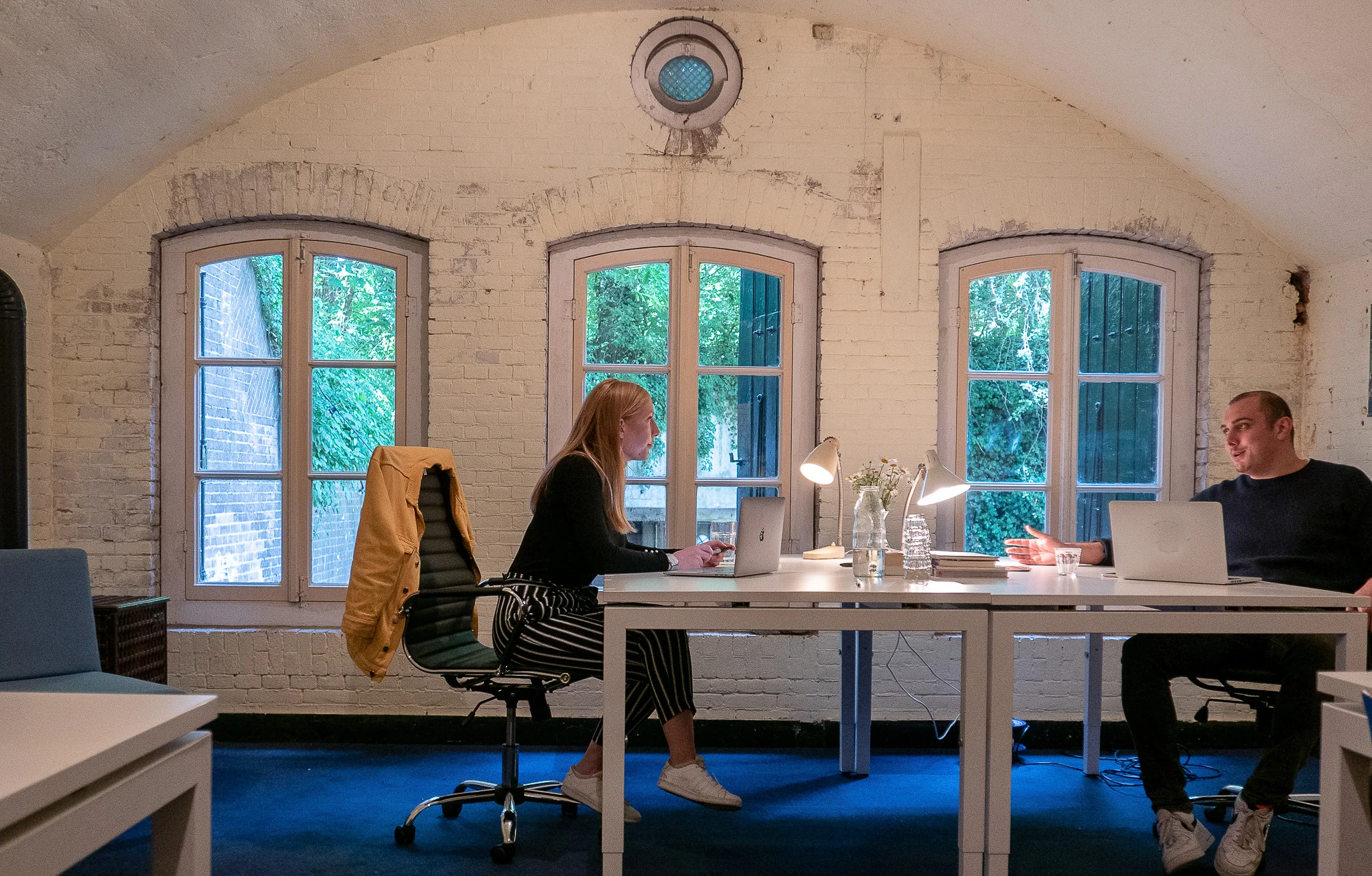 Twee mensen werken op laptops aan tegenovergestelde zijden van een bureau in een sfeervolle ruimte met gebogen plafond en grote ramen in Lekdijk.