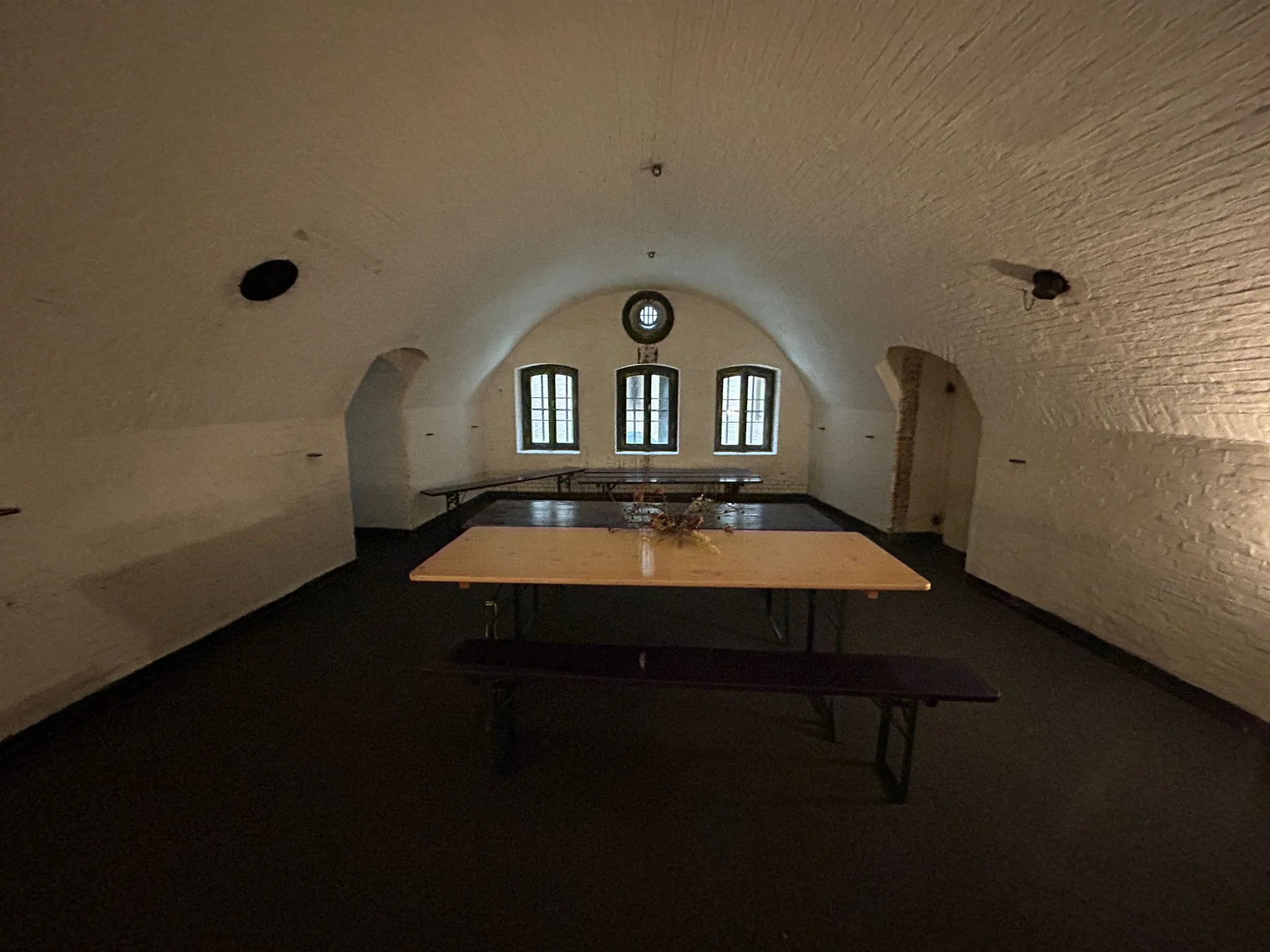 Een gewelfde kelderruimte met witte bakstenen muren, een houten tafel met banken en drie ramen aan de achterzijde.