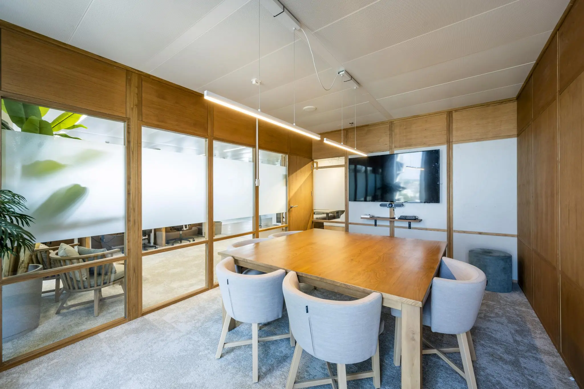 Vergaderruimte met houten tafel, zes stoelen, glazen wanden en een groot videoscherm aan de muur.