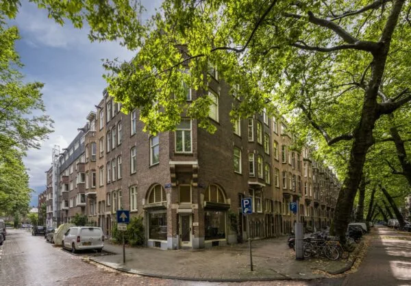Straathoek met klassieke bakstenen woongebouwen aan de Lomanstraat in Amsterdam, omringd door grote bomen en geparkeerde fietsen.