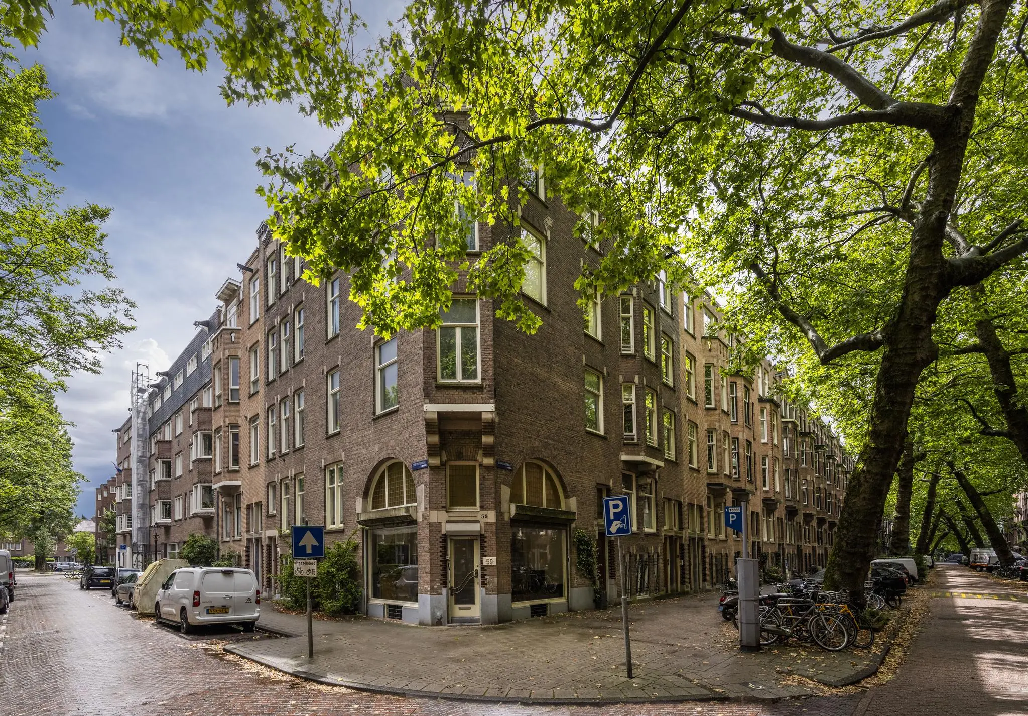 Straathoek met klassieke bakstenen woongebouwen aan de Lomanstraat in Amsterdam, omringd door grote bomen en geparkeerde fietsen.