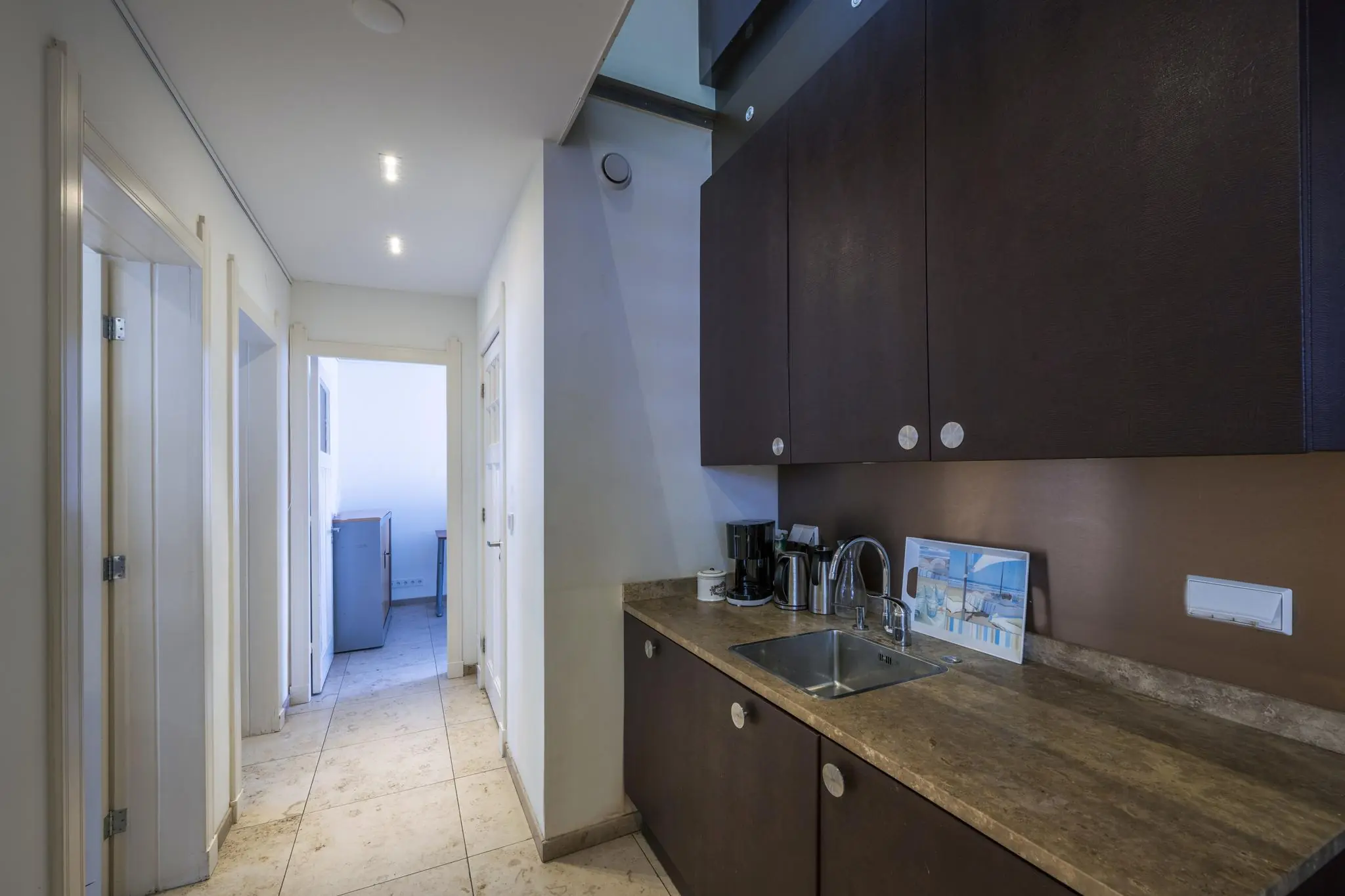 Moderne kitchenette aan de Lomanstraat met donkerbruine kasten, marmeren aanrecht en zicht op een gang met tegelvloer.