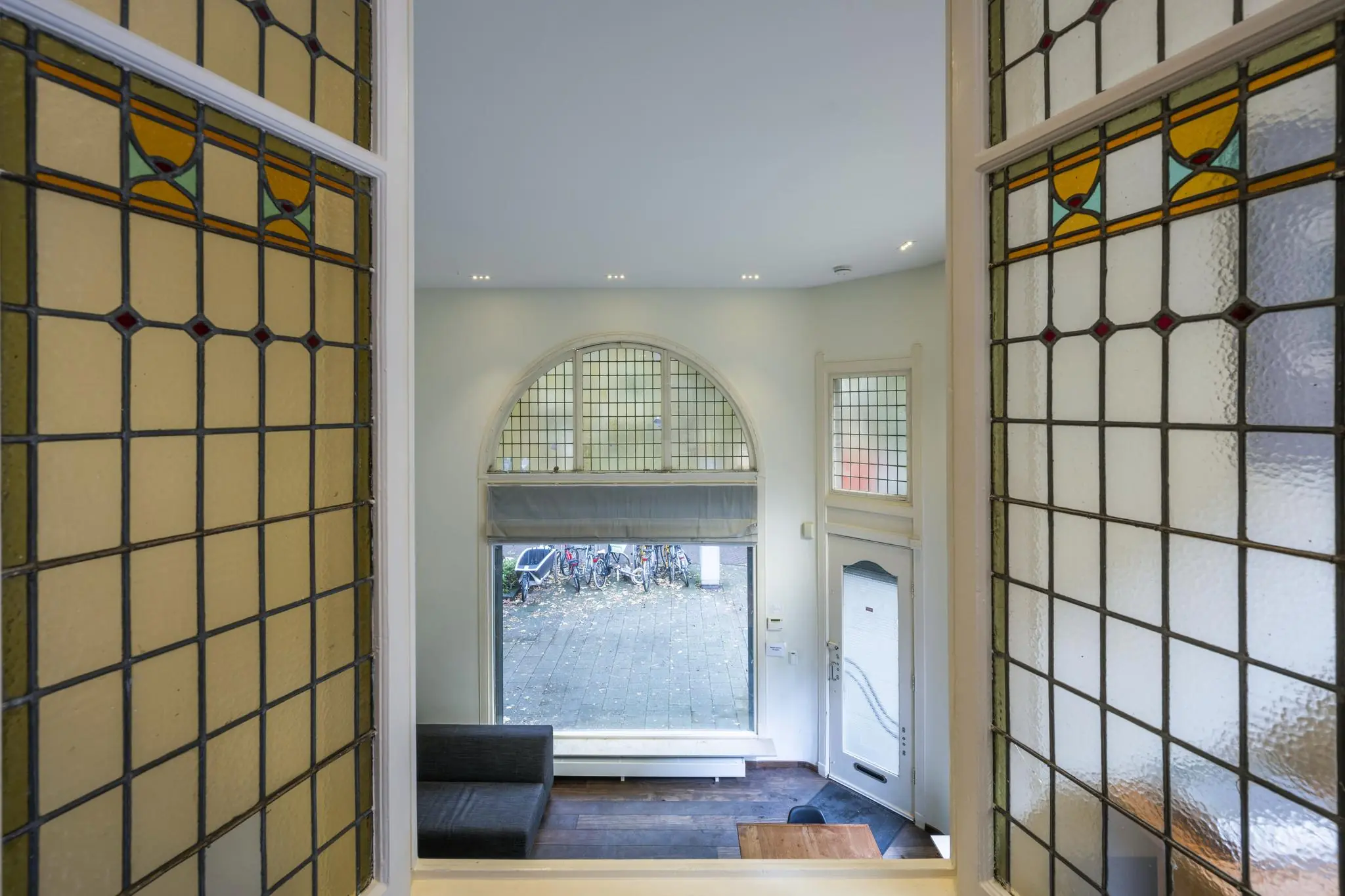 Interieur van een woonkamer aan de Lomanstraat met glas-in-loodramen en uitzicht op de straat met fietsen.
