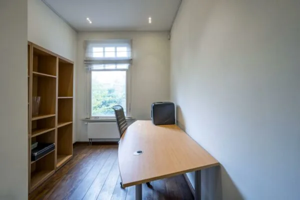 Kantoorruimte aan de Lomanstraat met houten vloer, bureau, bureaustoel, open kast en raam met uitzicht op groen.
